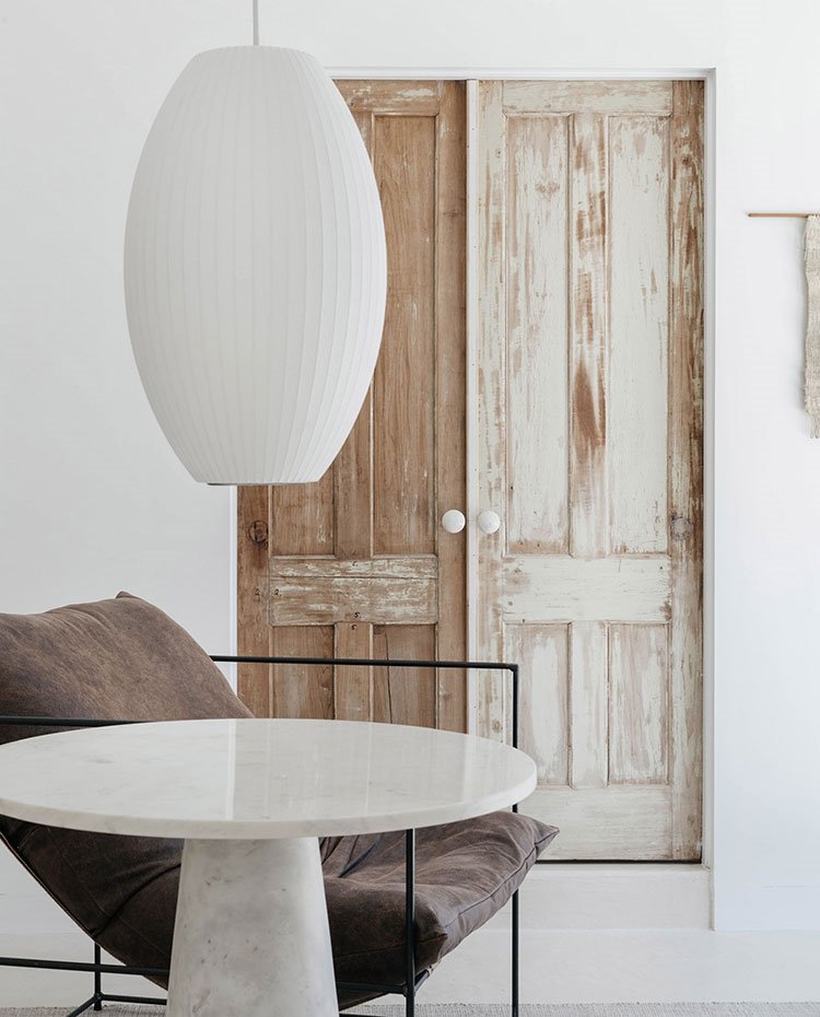 Luminaria suspendida blanca ovalada, mesa auxiliar blanca, butaca marrón con estructura tubular y puerta de madera decapada