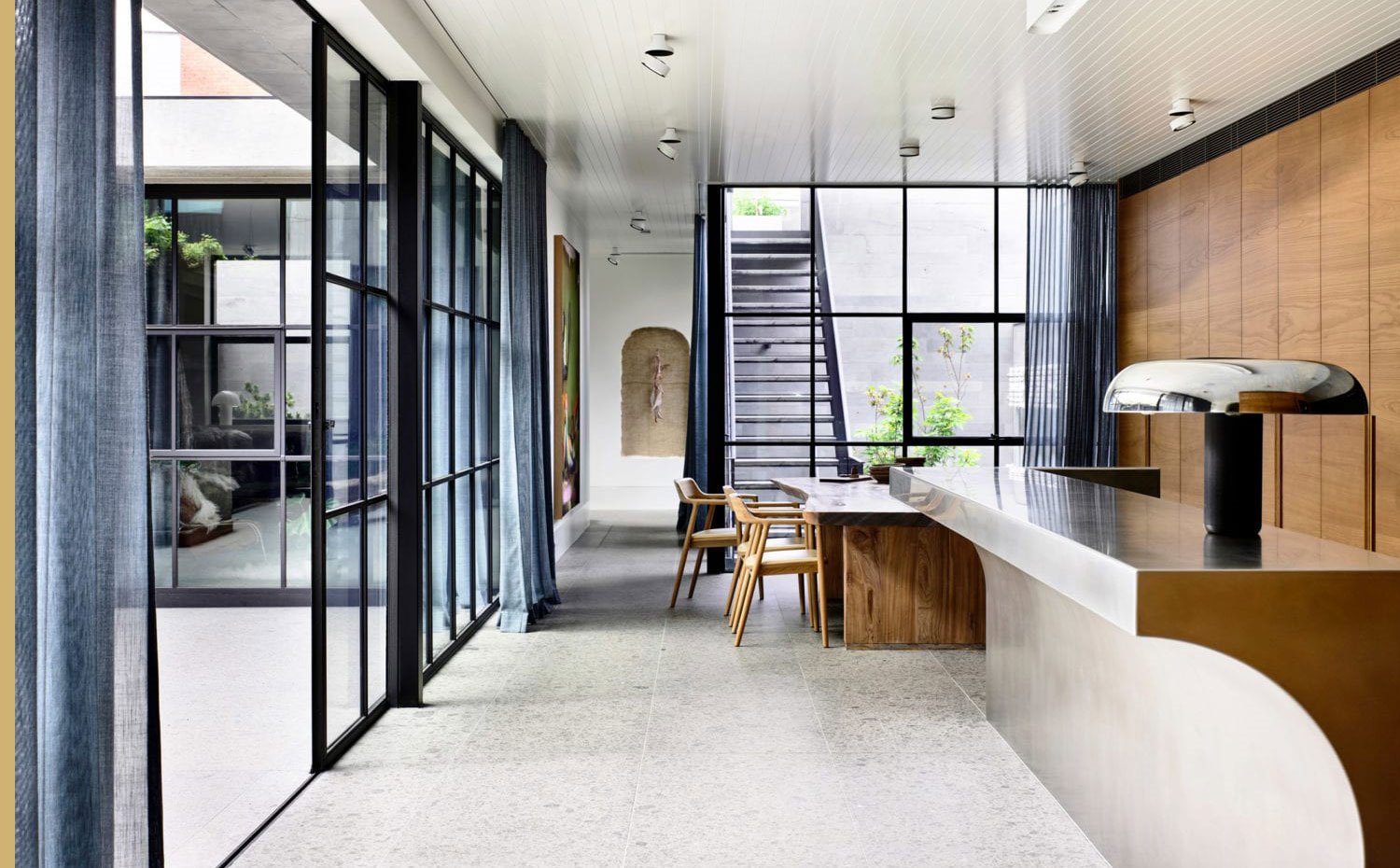 Cocina abierta con isla de acero, luminaria con pantalla cromada, mesa y sillas de office de madera, cerramiento acristalado 