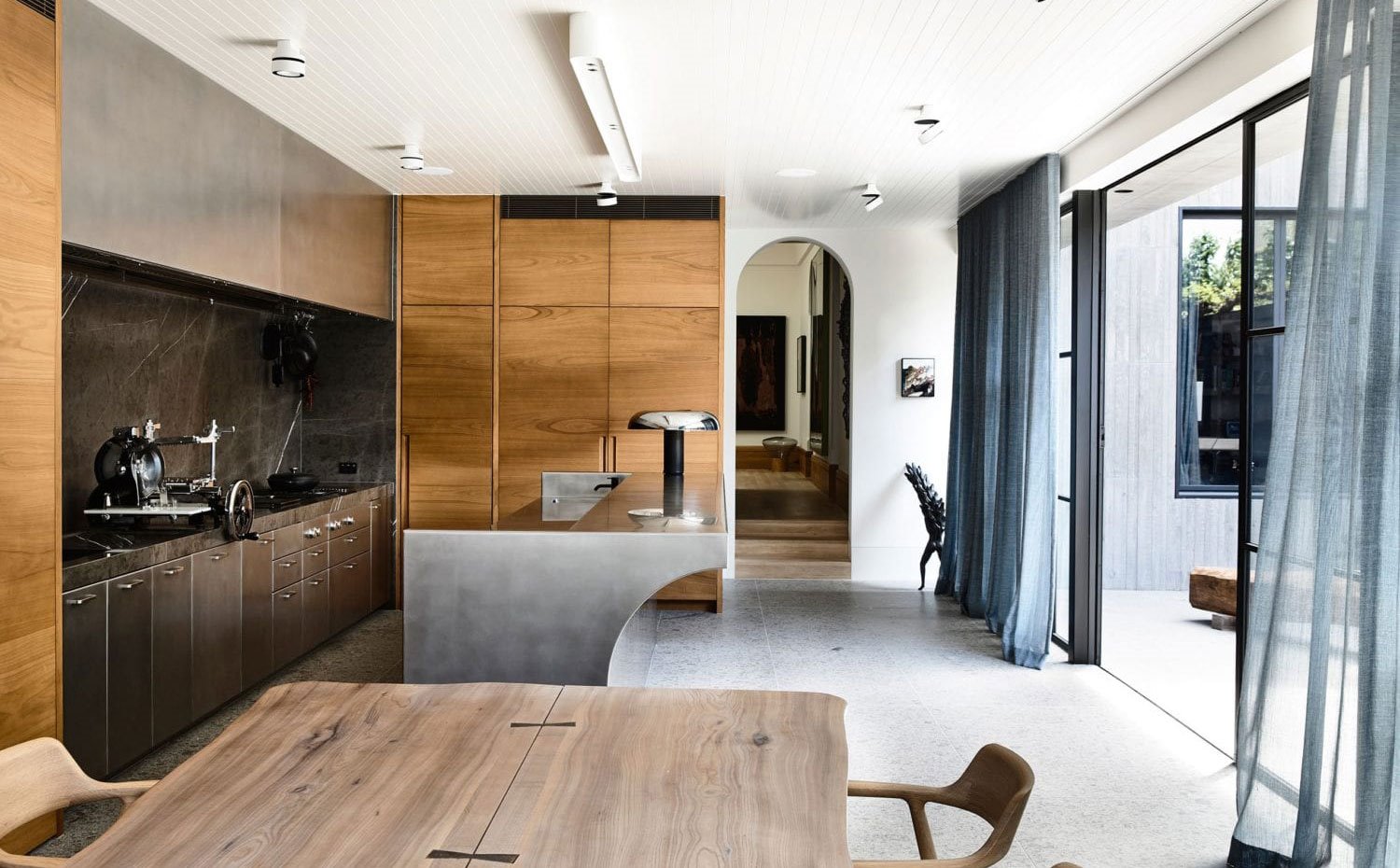 Cocina con mobiliario en madera y acero inoxidable, mesa y sillas de office de madera, cortinas azul grisáceo, cerramientos de cristal