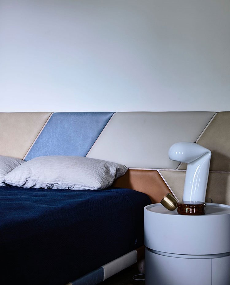 Dormitorio con cabecero tapizado en varios colores, mesilla circular en gris claro y luminaria de sobremesa  