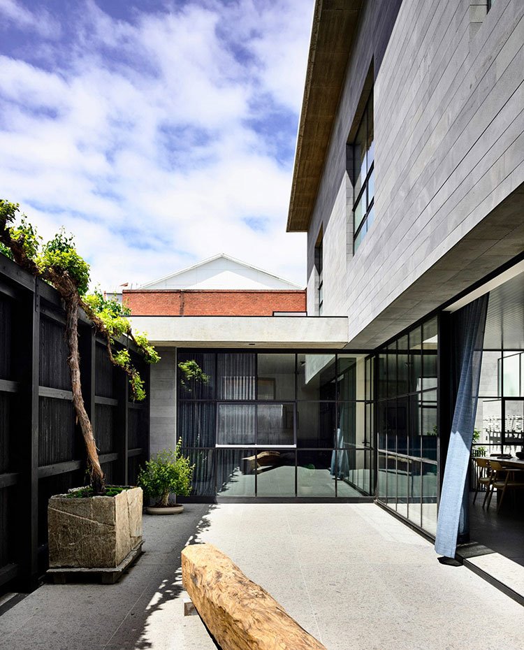 Patio interior vivienda con tronco de madera a modo de banco, árbol y planta y cerramiento en acero y cristal