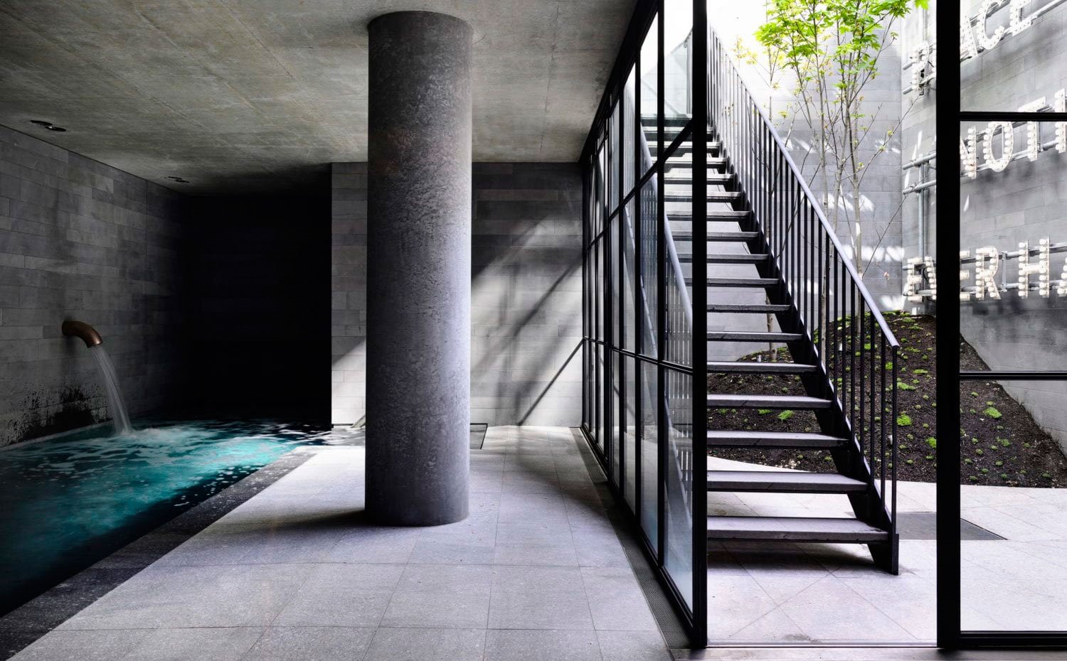 Piscina interior bajo estructura de hormigón, apertura a patio con cerramiento de cristal y escalera de acero en negro