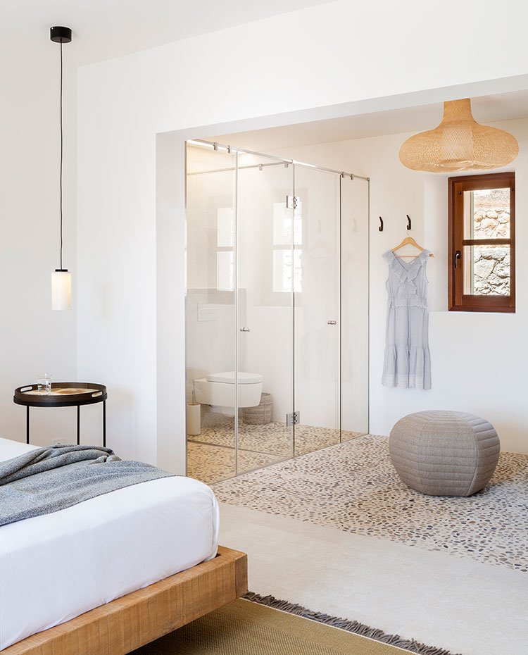 Dormitorio con baño semiabierto a base de cerramiento transparente, suelo de canto rodado y pouf grisáceo