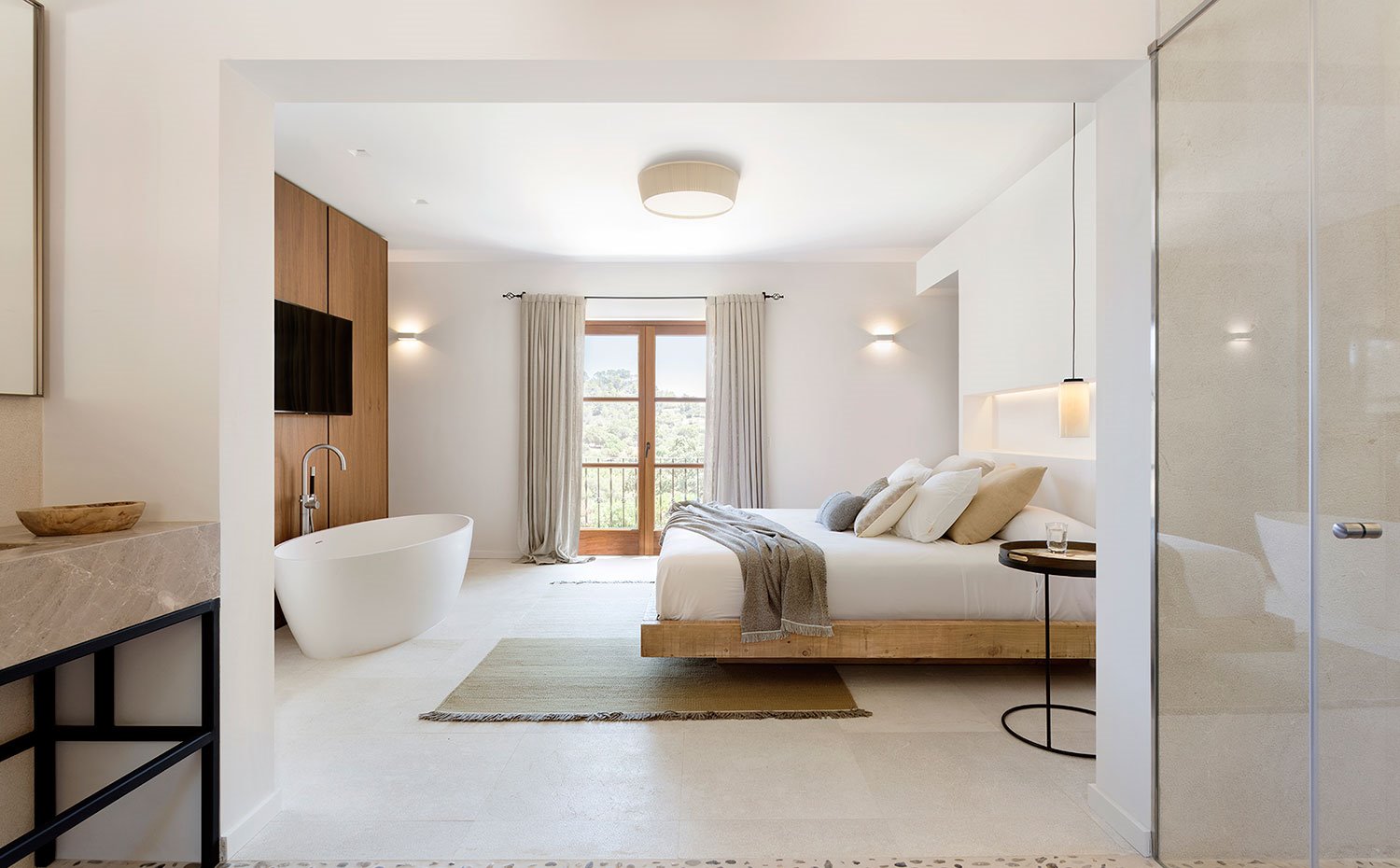 Dormitorio con cama en estructura de madera, mesa auxiliar negra cromada, baño abierto con encimera de piedra