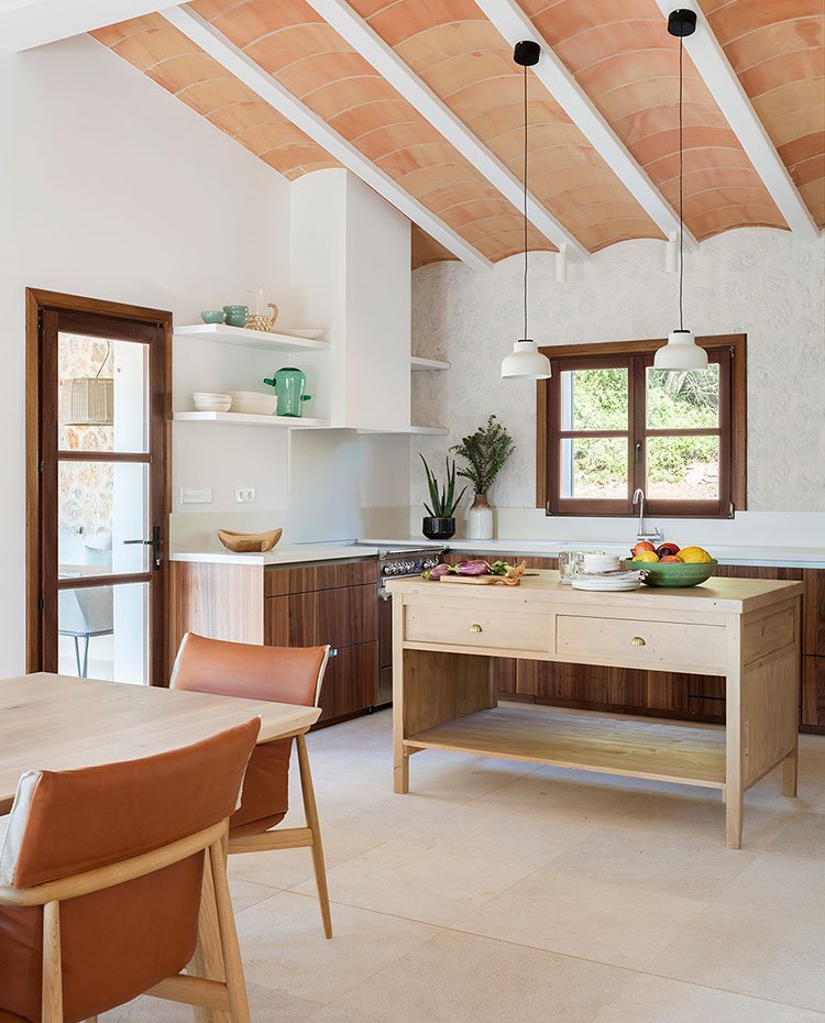 Mesas de piel y madera de comedor hacia cocina con isla de trabajo y luminarias blancas suspendidas