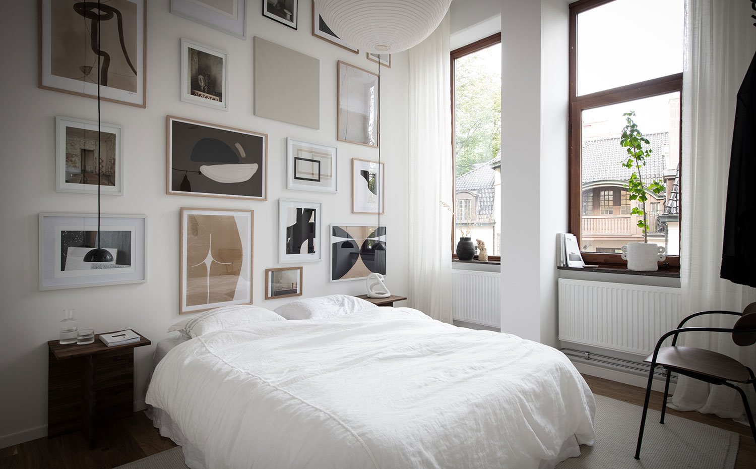 Dormitorio con frente de pared con láminas de diferentes tamaños, mesillas de madera, cortinas blancas