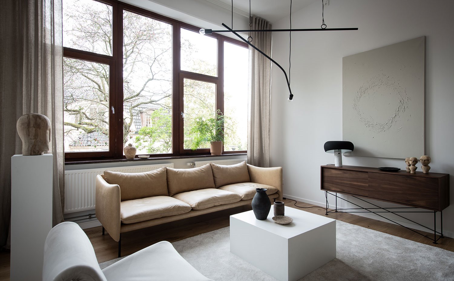 Salón con sofá beige de piel, pedestal blanco con escultura de madera, frente superior con cerramiento acristalado y perfilería en acabado madera