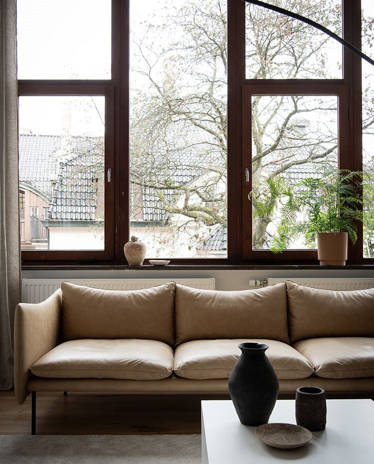 Salón con sofá beige de piel y repisa bajo ventana con jarrones y plantas