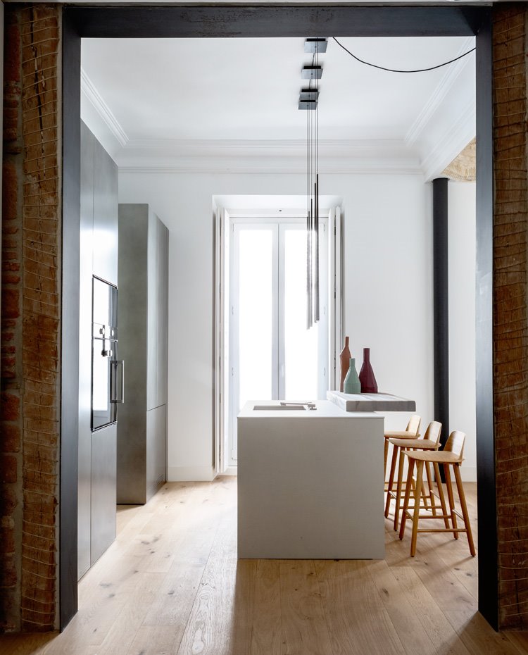 Cocina con mobiliario gris, isla de trabajo y balda de madera a modo de barra de office con taburetes de madera