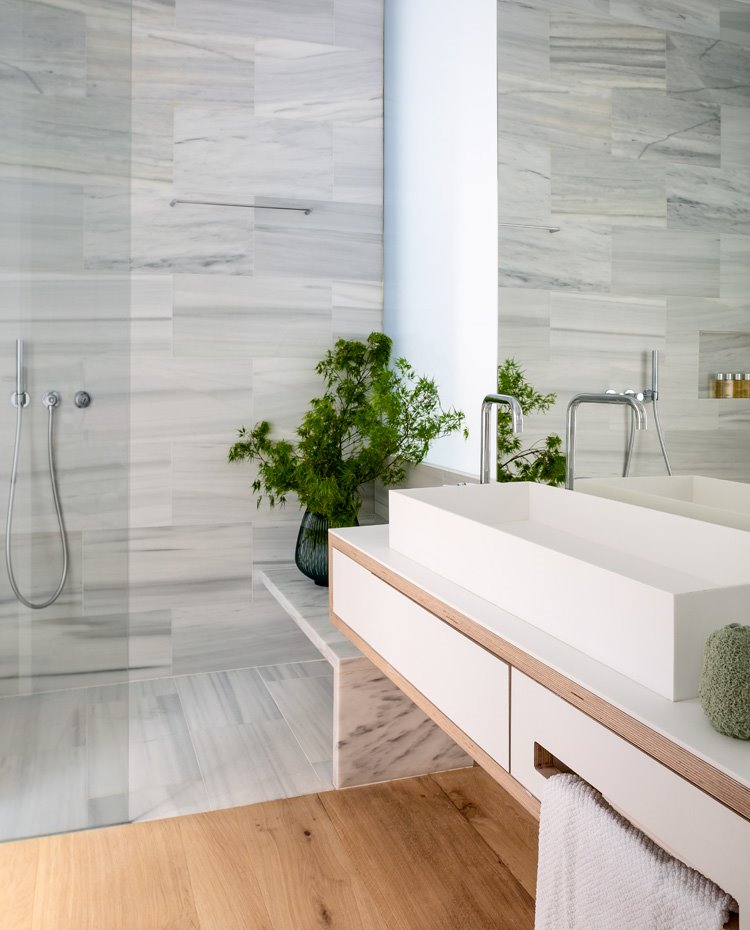 Cuarto de baño con suelo y revestimiento en marmol con tabiquería de cristal, módulo lavabo suspendido en blanco y madera