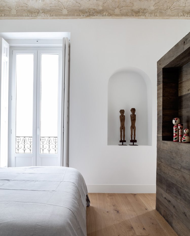 Dormitorio con módulo separador en madera oscura, hornacina en pared con esculturas, paredes en blanco con detalles originarios en techo, balcón a exterior con pórticos de madera blanca 