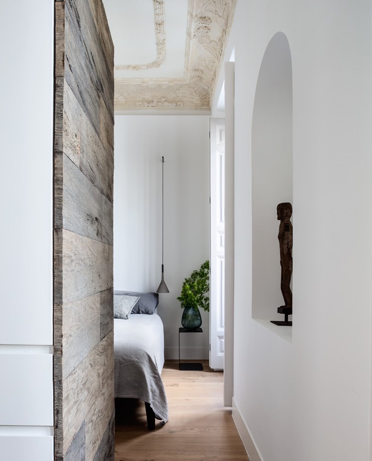 Luminaria suspendida con pantalla en gris, mesita de noche de estructura metálica en forma de C, revestimiento de armarios en blanco y madera natural sin tratar