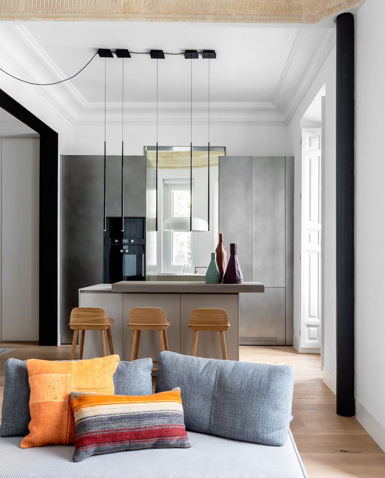 Salón y cocina con luminarias suspendidas sobre barra de madera de office, taburetes de madera hacia isla con mobiliario gris, molduras en techos