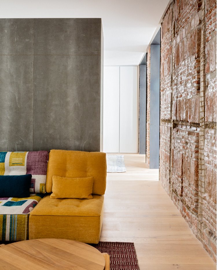 Sofá modular en color mostaza y a cuadros malva y verde, mesa de madera, miro de hormigón y pared con ladrillo de obra vista
