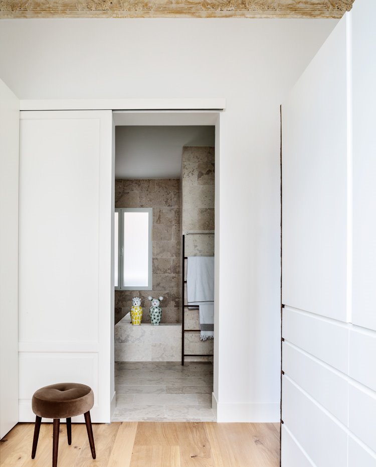 Zona vestidor con armarios lacados en blanco, cuarto de baño revestido de piedra tras puerta corredera