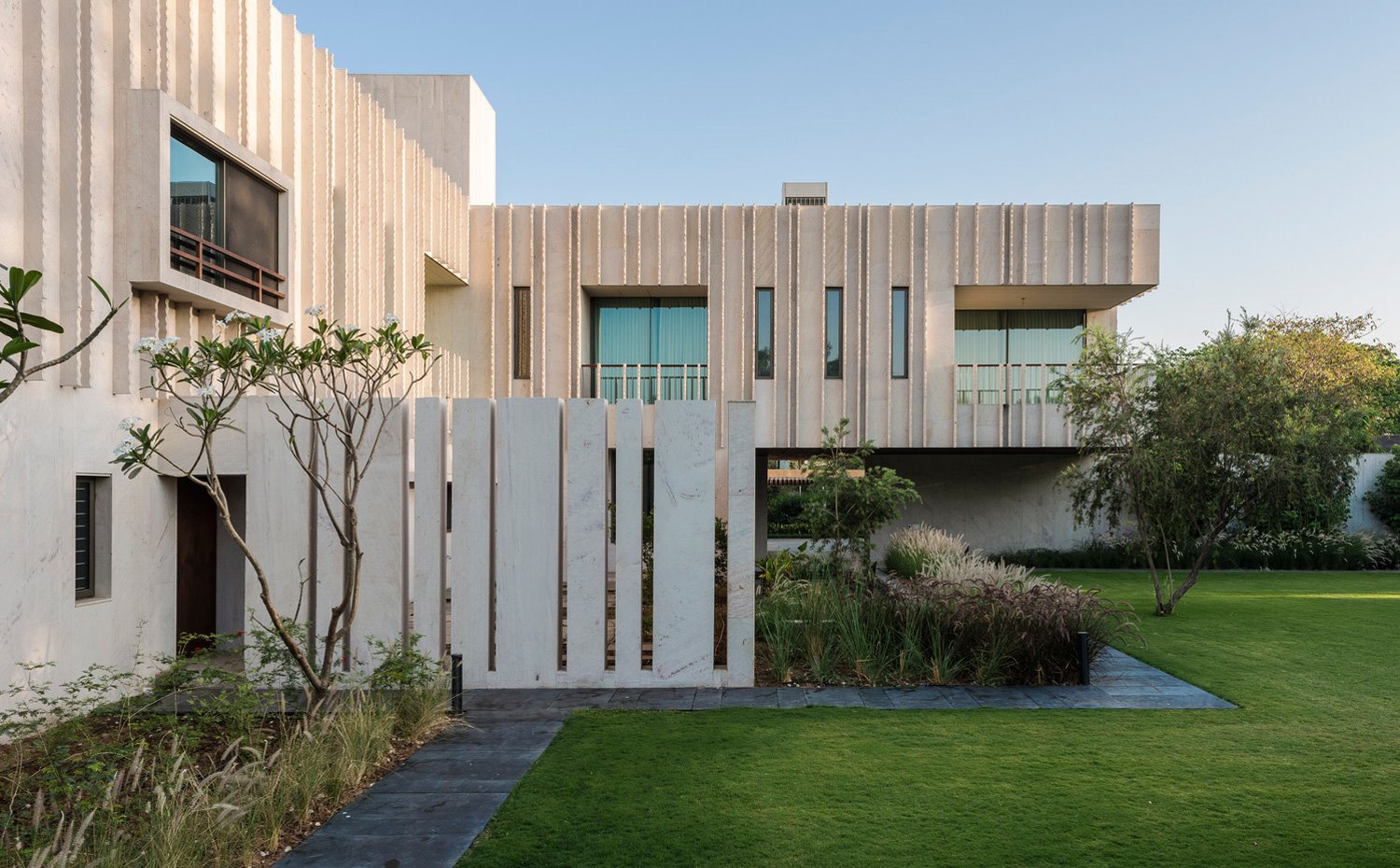 Exterior vivienda con fachada texturaizada, árboles y arbustos delimitando el perímetro