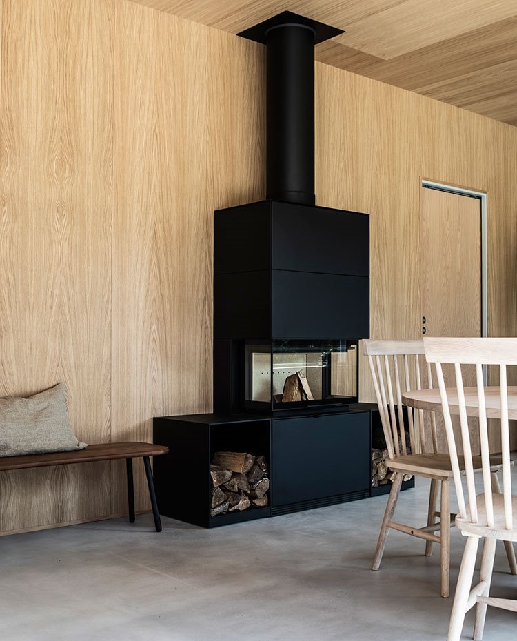Chimenea negra de forma resctangular, con zona de almacenaje para leña, bancada de madera oscura y mesa y sillas de comedor