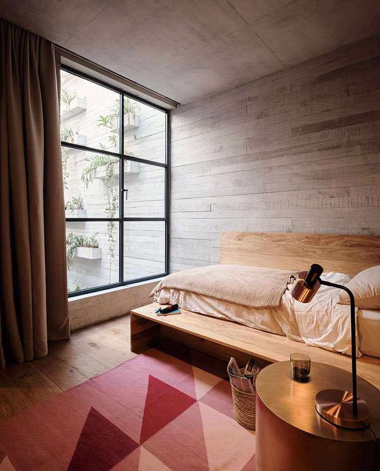 Dormitorio secundario con estructura de madera, mesilla circular con sobre dorado, luminaria de sobremesa a juego, alfombra en tonos rosados, gran apertura a patio interior