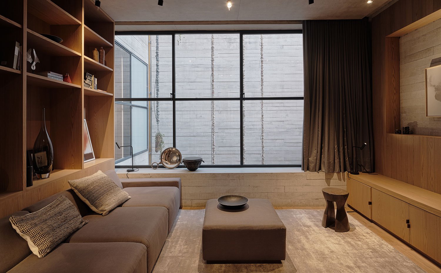 Zona de estar con sofá y pouf central en marrón, mobiliario en madera clara, alfombra en tonos tierra, taburete de madera