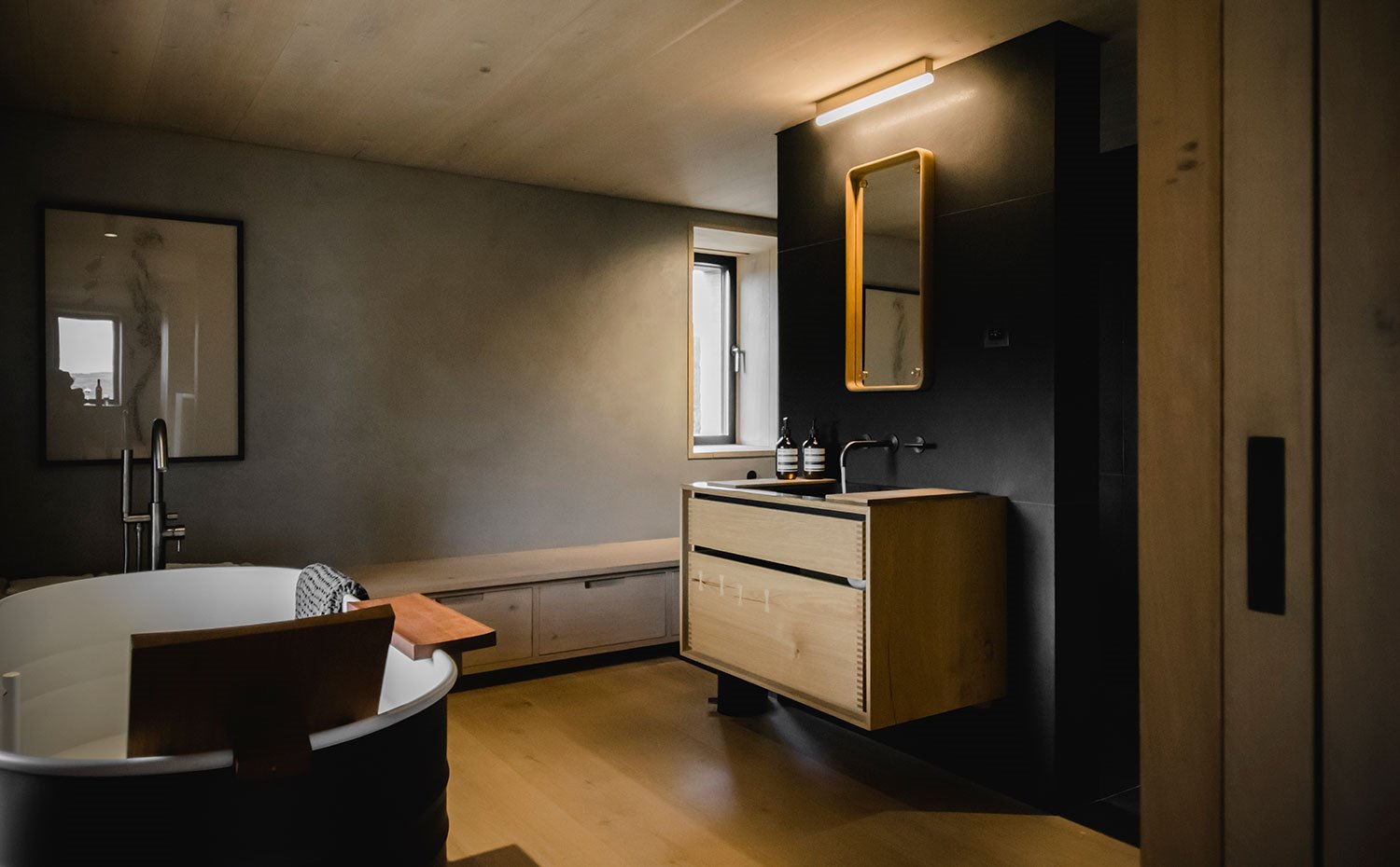 Cuarto de baño con bañera en blanco y negro, mobiliario de madera en zona de lavamanos, módulos bajos en pared