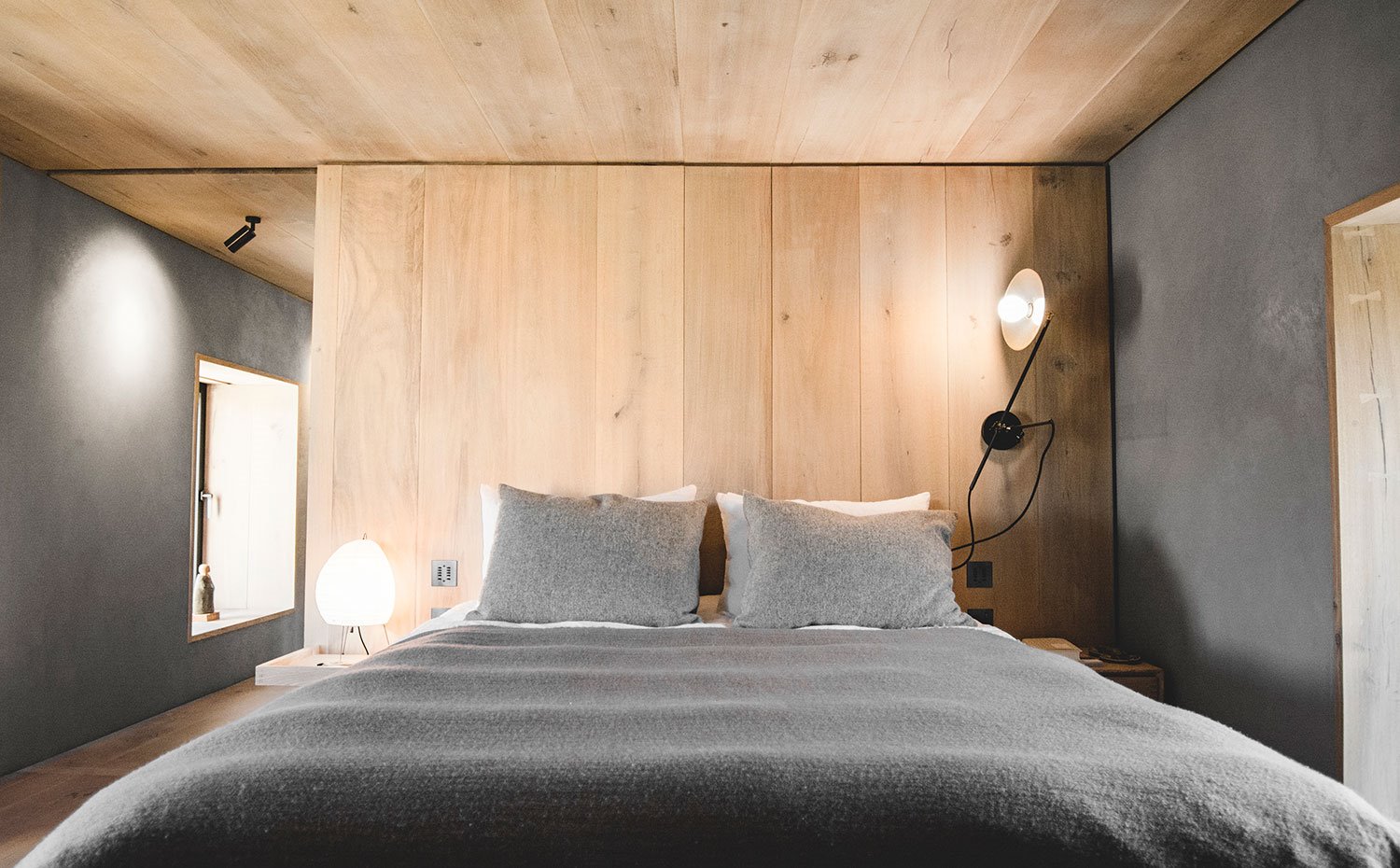 Dormitorio con cabecero de madera en acabado natural, pared estucado veneciano en gris, apliques negros