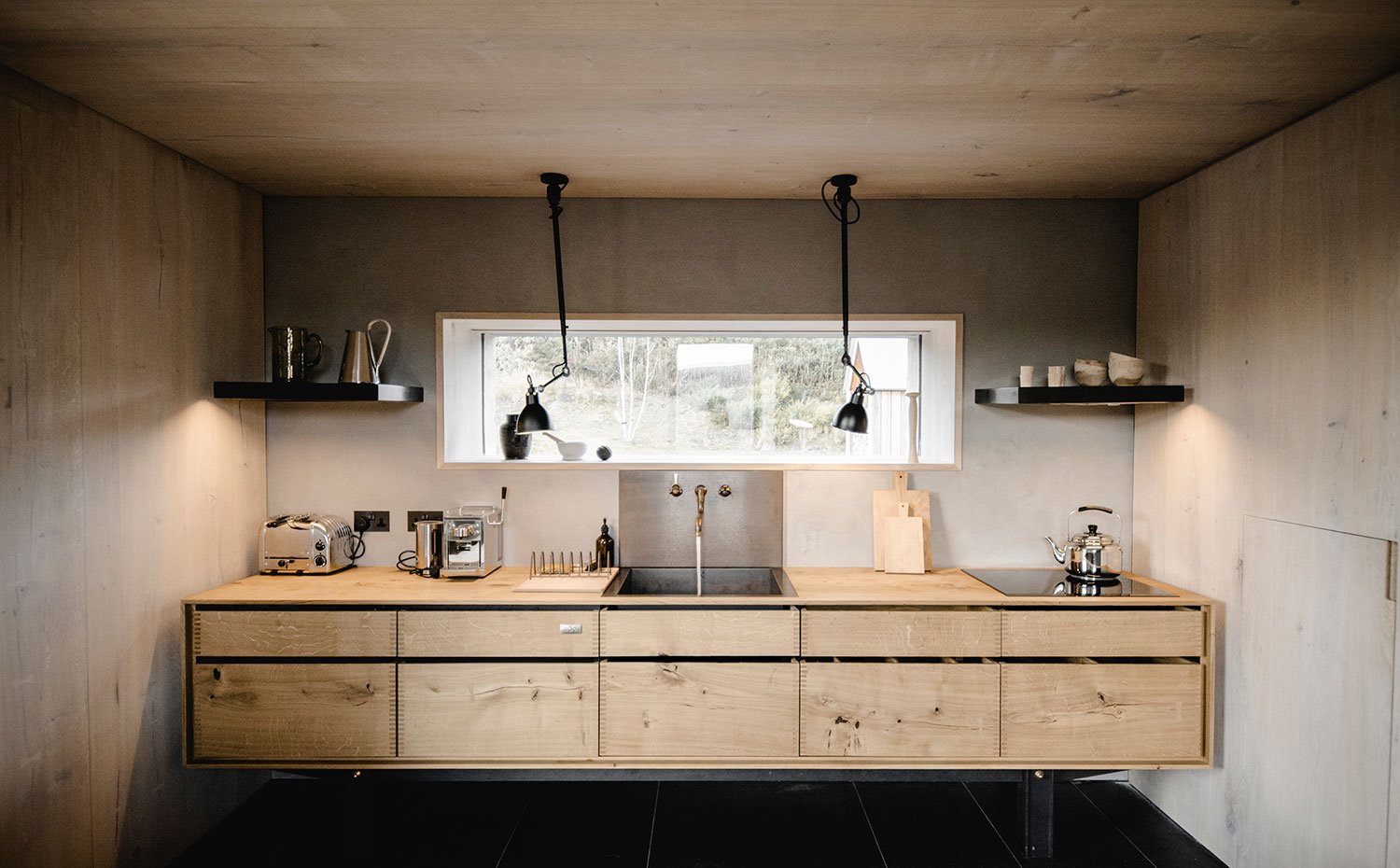 Frente de cocina, con módulos bajos suspendidos de madera natural, electrodomésticos cromados sobre encimera