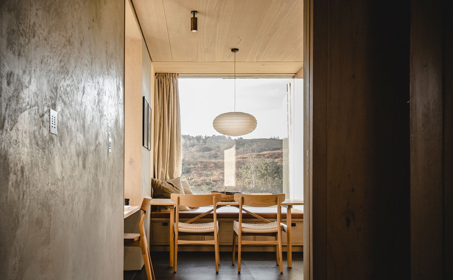 Zona comedor con sillas de madera, luminaria suspendida con pantalla ovalada, foco negro integrado en techo laminado en madera