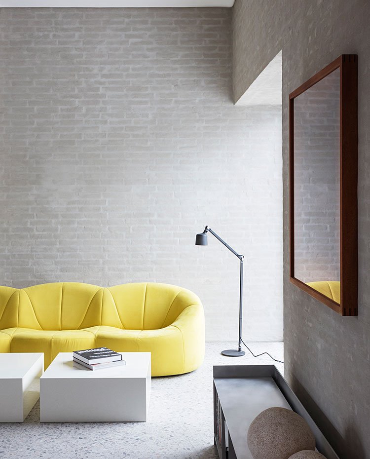 sofá en amarillo, luminaria de pien en negro, mesas simétricas en blanco, pared de ladrillo y hormigón