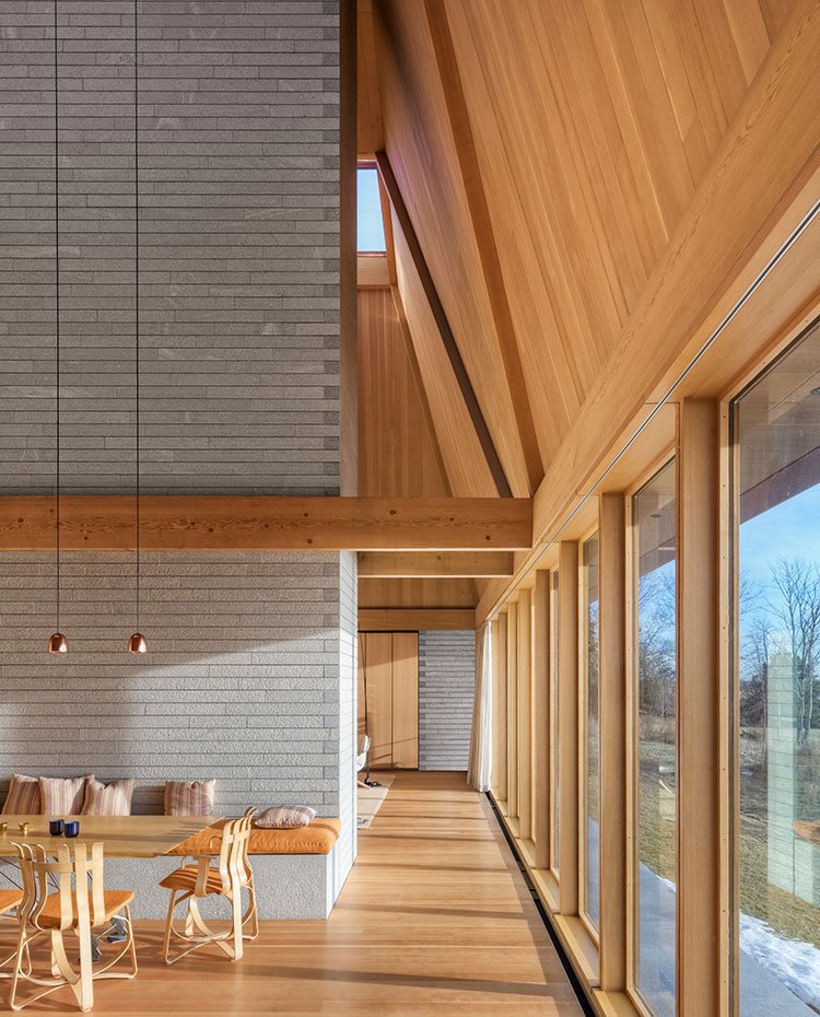 Revestimiento de piedra y madera, cerramiento acristalado, mesa de comedor con luminarias suspendidas a gran altura, banco y sillas de madera