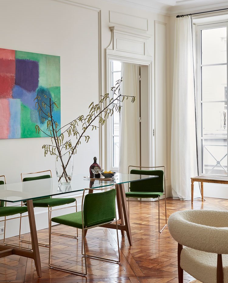 Comedor con mesa con sobre de cristal y sillas verdes, suelo de madera, puerta con molduras, butacas en blanco