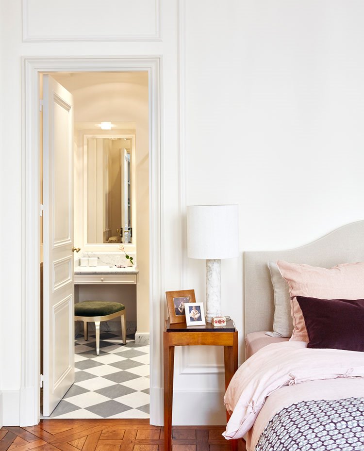 Dormitorio con mesilla de madera, luminaria de sobremensa en blanco, puerta a cuarto de baño
