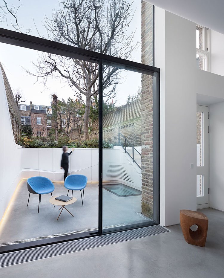Cerramientos de cristal en la parte frontal de la vivienda con terraza de planta irregular y de perímetro ondulante con sillas en azul