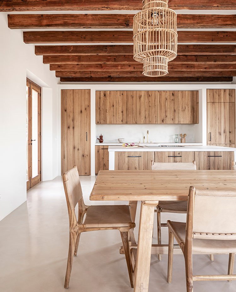 Mesa de comedor y sillas, todo de madera, junto a cocina, iuminaria suspendida de formas ligeras, vigas en techo