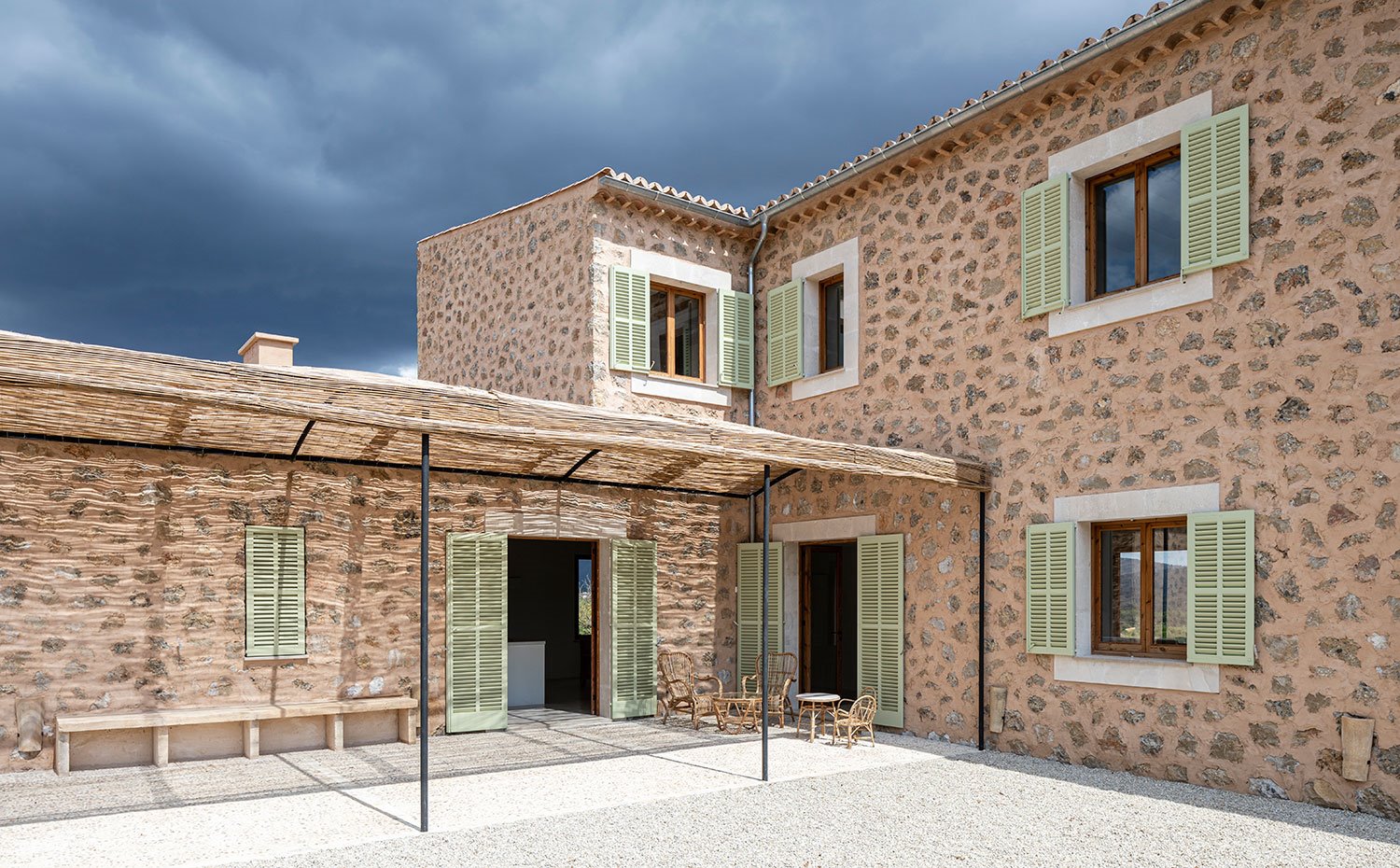 Porche exterior con estructura de acero y fibra natural protectora, contraventanas verdes, fachada en piedra