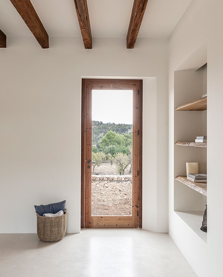 Puerta con perfilería de madera a exterior, baldas de madera en hueco integrado en pared, cesto de mimbre con cojines