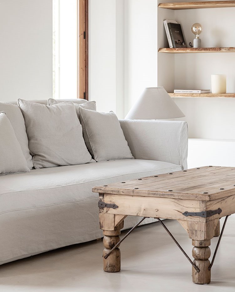 Sofá y cojines a juego en gris claro, mesa de centro de madera antigua, baldas en hueco de pared en madera, perfilería de cerramientos también de madera