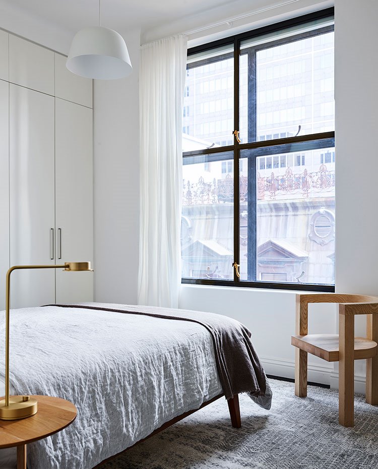 Dormitorio con silla y mesillas de noche de madera, luminaria de sobremesa en acabado latón, ventanas de doble cristal hacia exterior, amarios encastrados en blanco