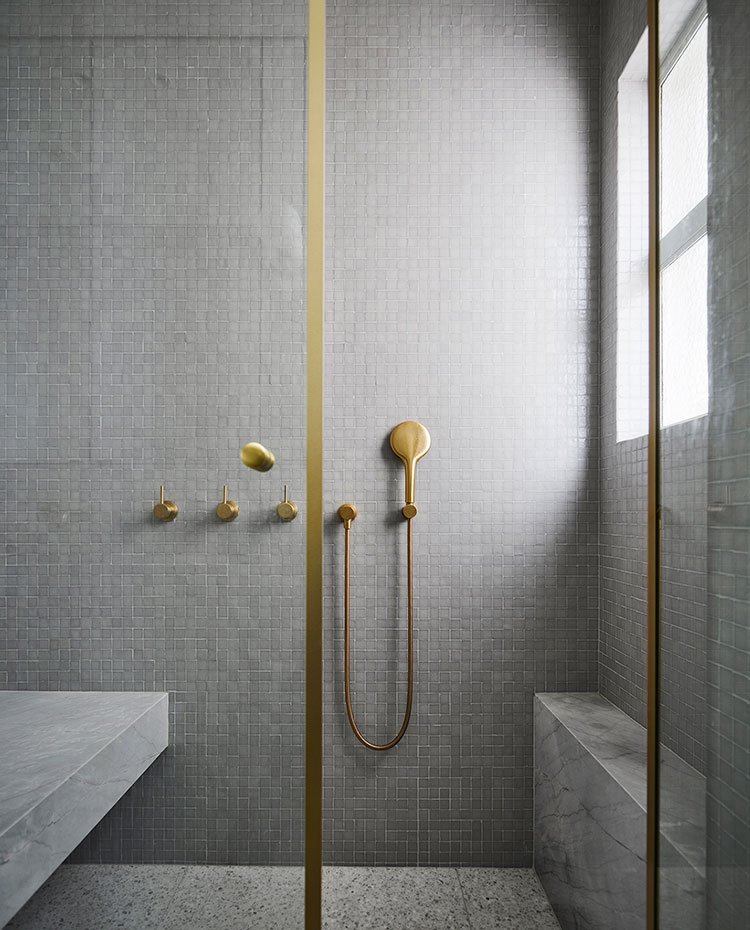 Ducha de cuarto de bajo con mampara de cristal transparente y estructura de obra, revestimiento el porcelánico gris y grifería y perfilería en acabado latón
