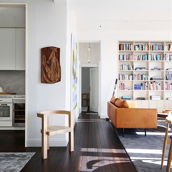 Salón con librería a pared sobre mueble bajo, sofá de color ocre, alfombra gris sobre suelo de madera, silla de madera, acceso a cocina