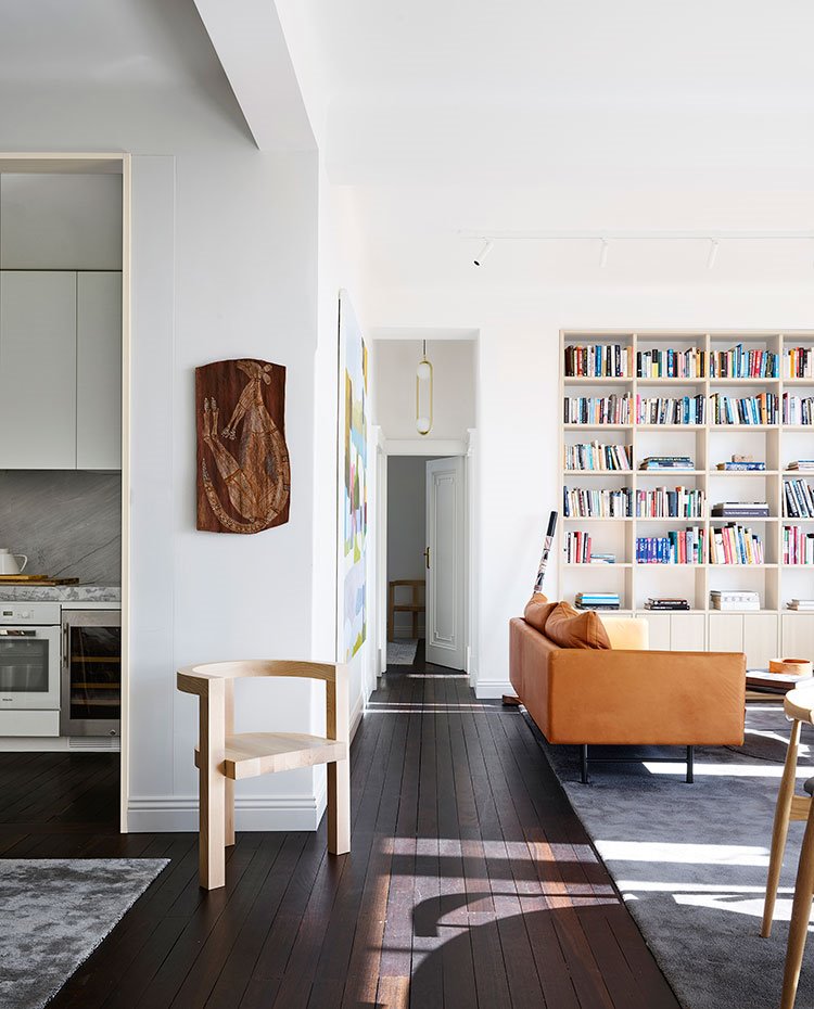 Salón con librería a pared sobre mueble bajo, sofá de color ocre, alfombra gris sobre suelo de madera, silla de madera, acceso a cocina