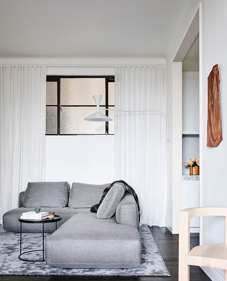 Sofá en forma de L frente a cocina, mesita auxiliar negra circular y luminaria a modo de aplique en blanco frente a cortinas blancas