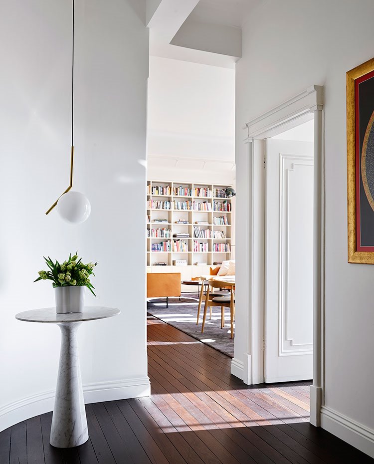 Zona de maso con moldura en marco puerta y zócalos, suelo de madera, iibrería en salón, mesita auxiliar con planta y luminaria suspendida con esfera blanca
