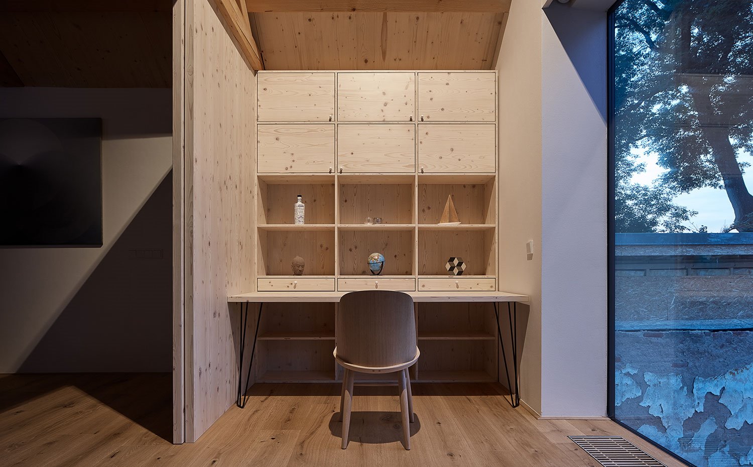 Zona de trabajo con mesas, silla y estantería de madera junto a apertura exterior de cristal.transparente