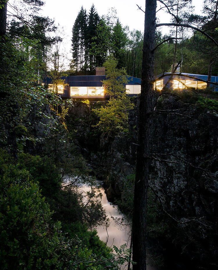 Vista frontal de la fachada con el interior iluminado, entorno natural y agua en cascada