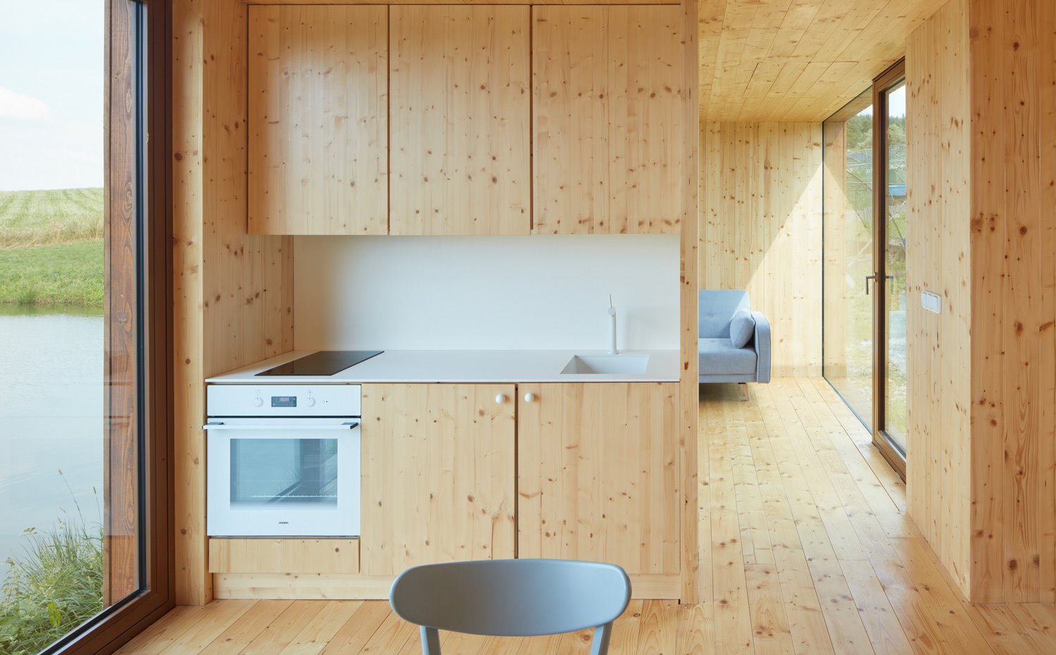 Cocina revestida de madera, con encimera y salpicadero en blanco, sillas y sofá en grisl, aperturas laterales a exterior