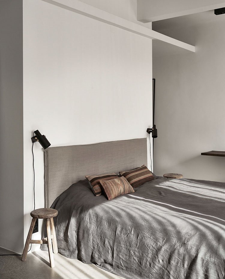 Dormitorio principal con cabecero de cama en lino, apliques de foco negro y taburete de madera