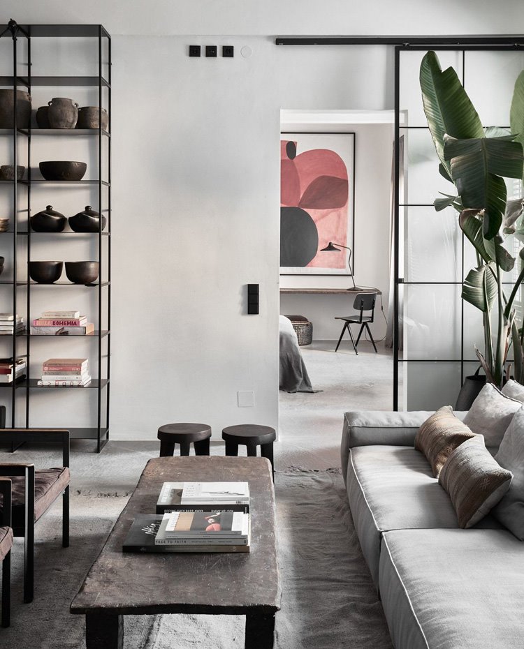 Zona de salón con mesa de centro rectangular en madera, sofá en gris claro, taburetes a juego de madera, estantería de hacero