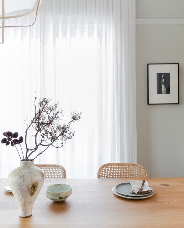 Jarrón y menaje en porcelana texturizada sobre mesa de comedor de madera, paredes pintadas en gris claro y cortinas en blanco