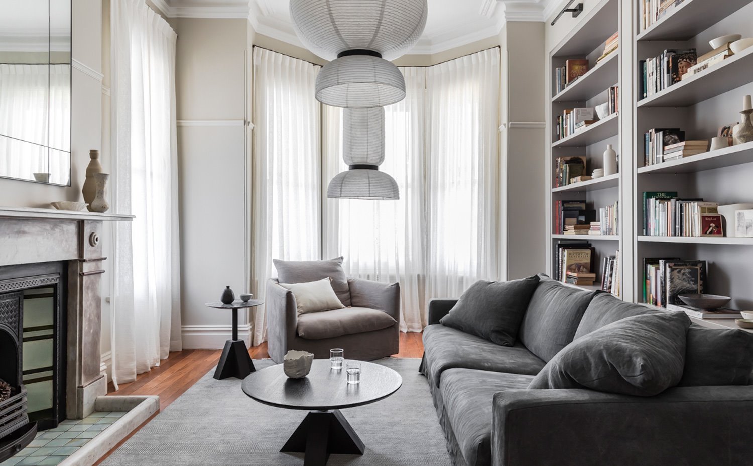 Salón con chimenea y librería a medida, sofá en gris y butaca en tonos tierra, mesa de centro redonda y luminarias suspendidas