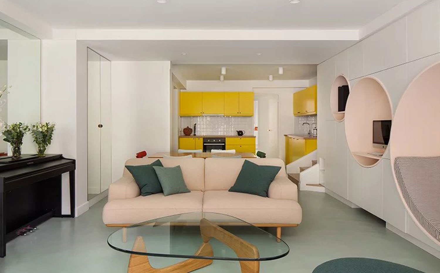 Salón con cocina al fondo, sofá rosa con cojines en verde, mesa de centro de cristal y madera, piano y armario con frentes de espejo.
